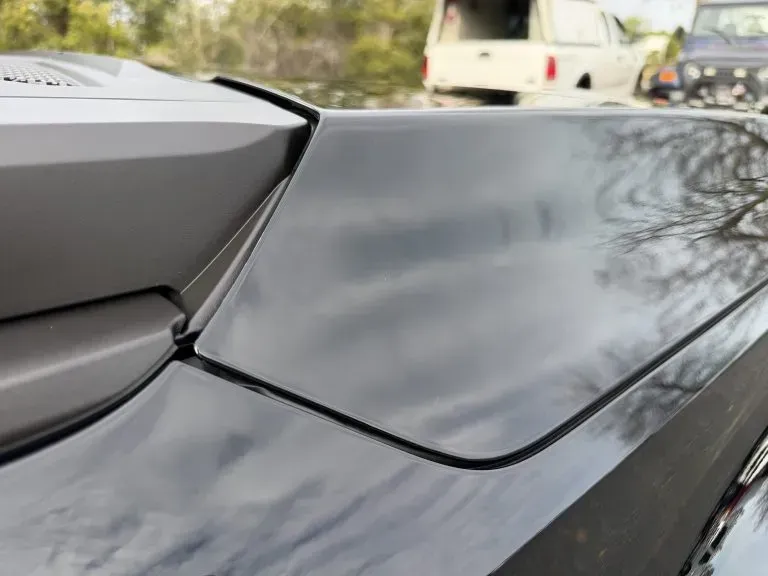 Close-up of a black car’s body panel with cloudy reflections and a blurred white van in the background