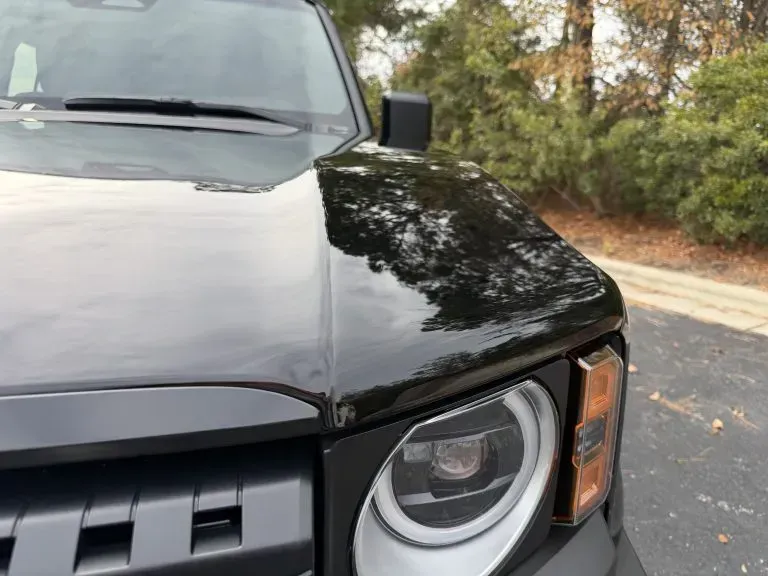 Close-up of a black SUV’s front corner with a headlight and hood reflecting trees.