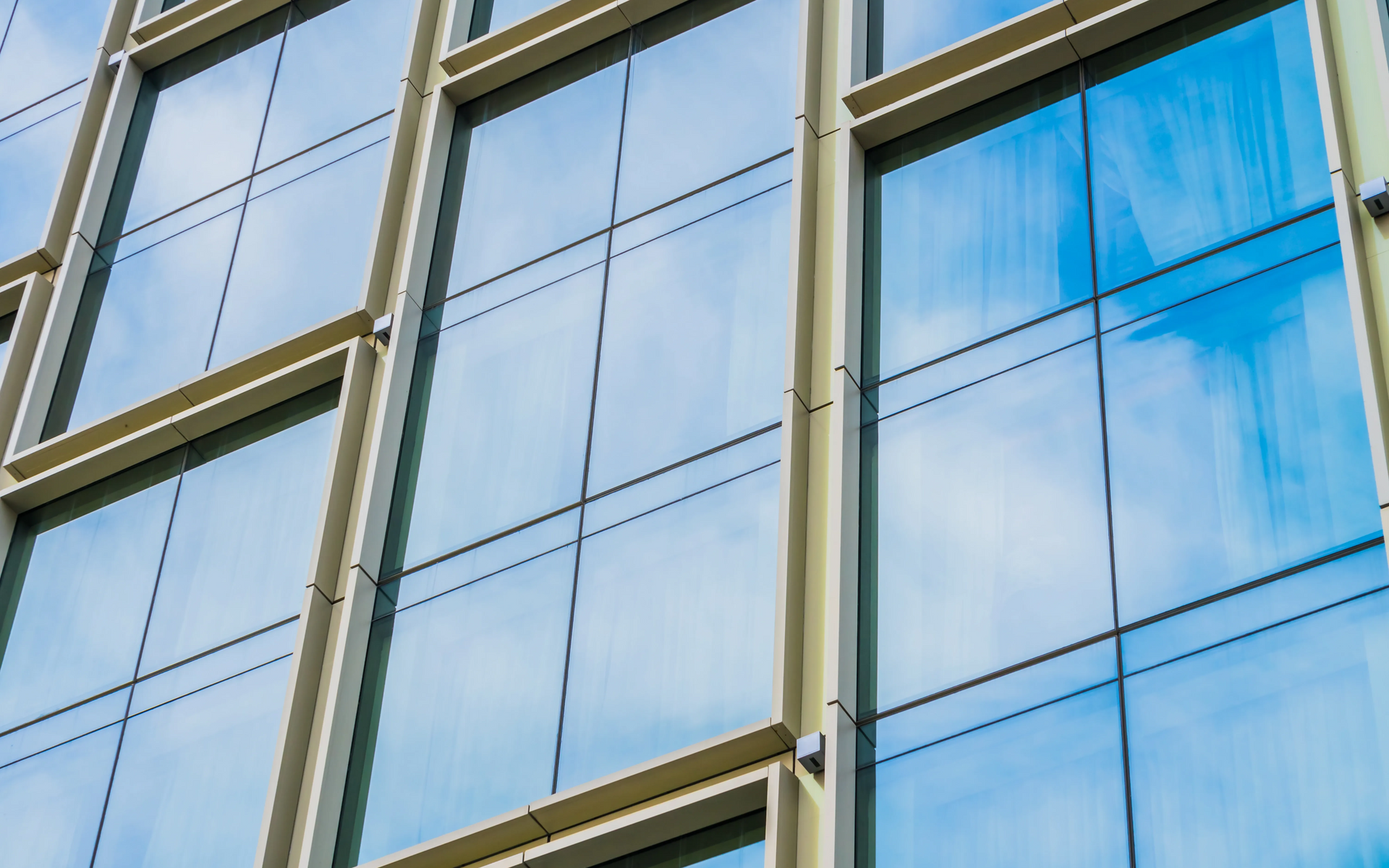 Glass office building facade with beige framing and blue-tinted windows reflecting clouds