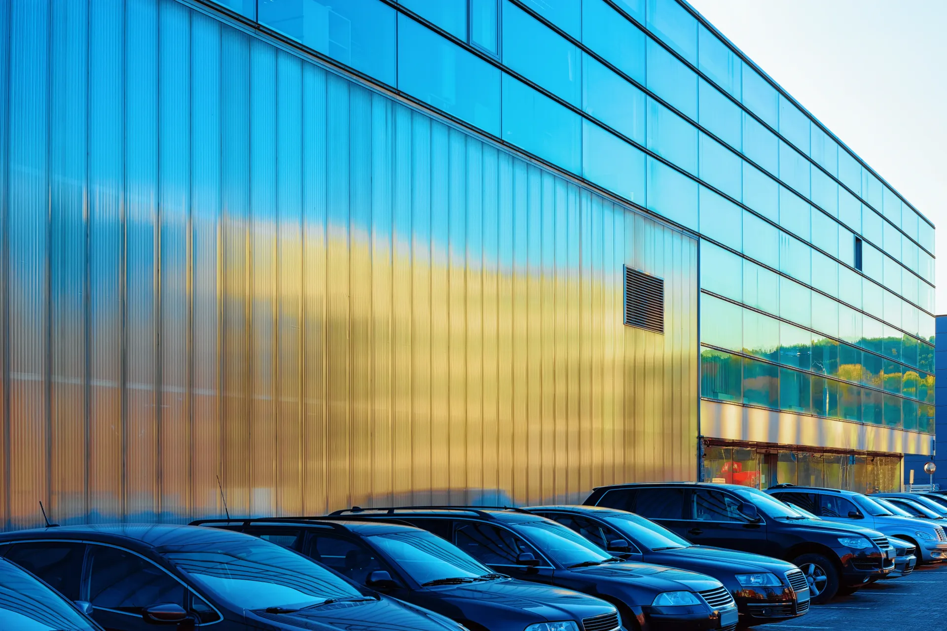 Modern glass building with blue-tinted windows and rows of parked cars in front