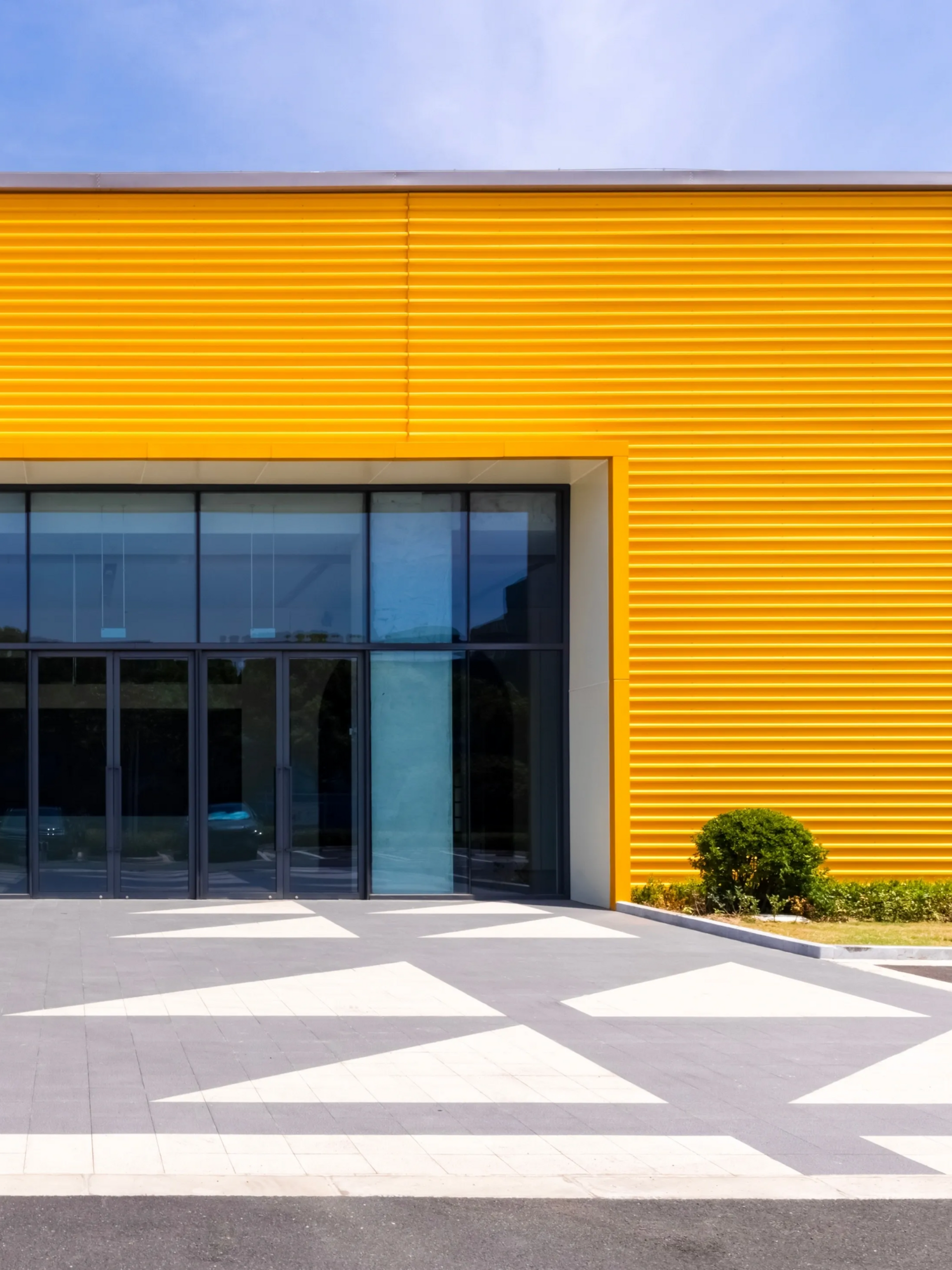 Modern building entrance with bright yellow facade and large glass doors, viewed from a geometric paved courtyard