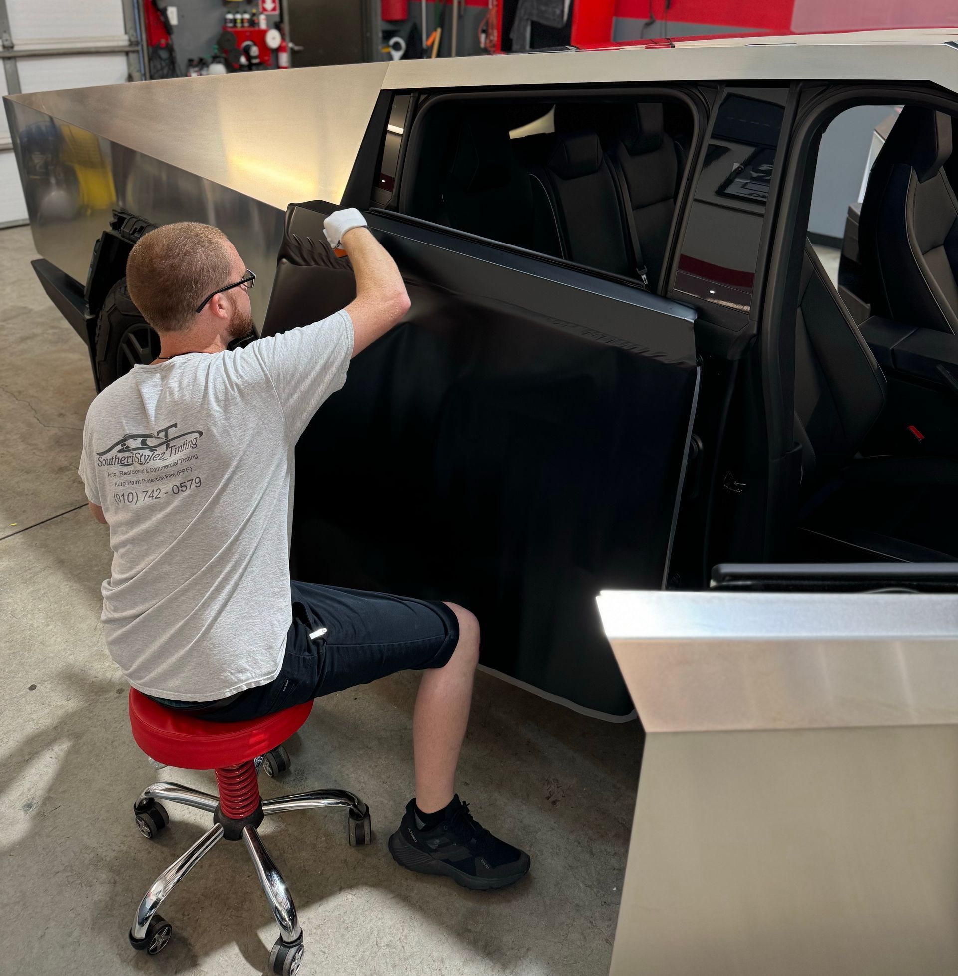 Man kneels beside a black off-road trailer, inspecting a side panel in a garage.