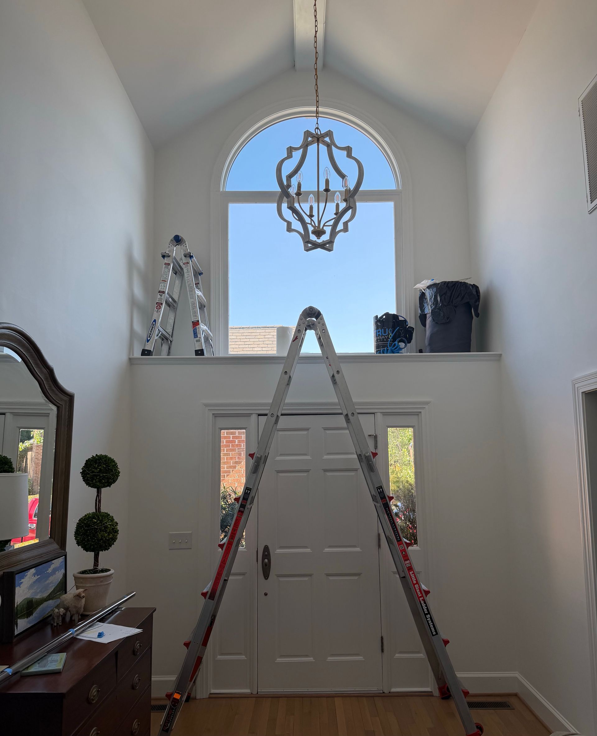 Tall foyer with high arched window, chandelier, and a ladder standing in front of double doors.