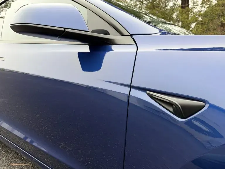 Blue car side mirror and door close-up with chrome trim and a triangular vent badge.