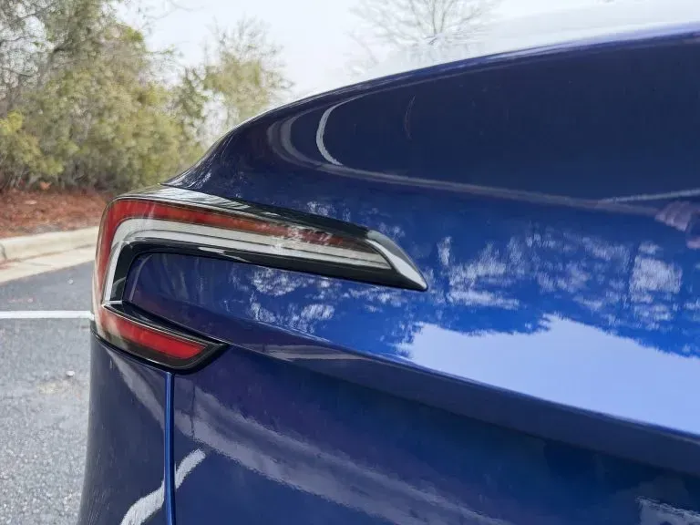 Close-up of a blue car’s rear taillight and body panel in a parking lot.