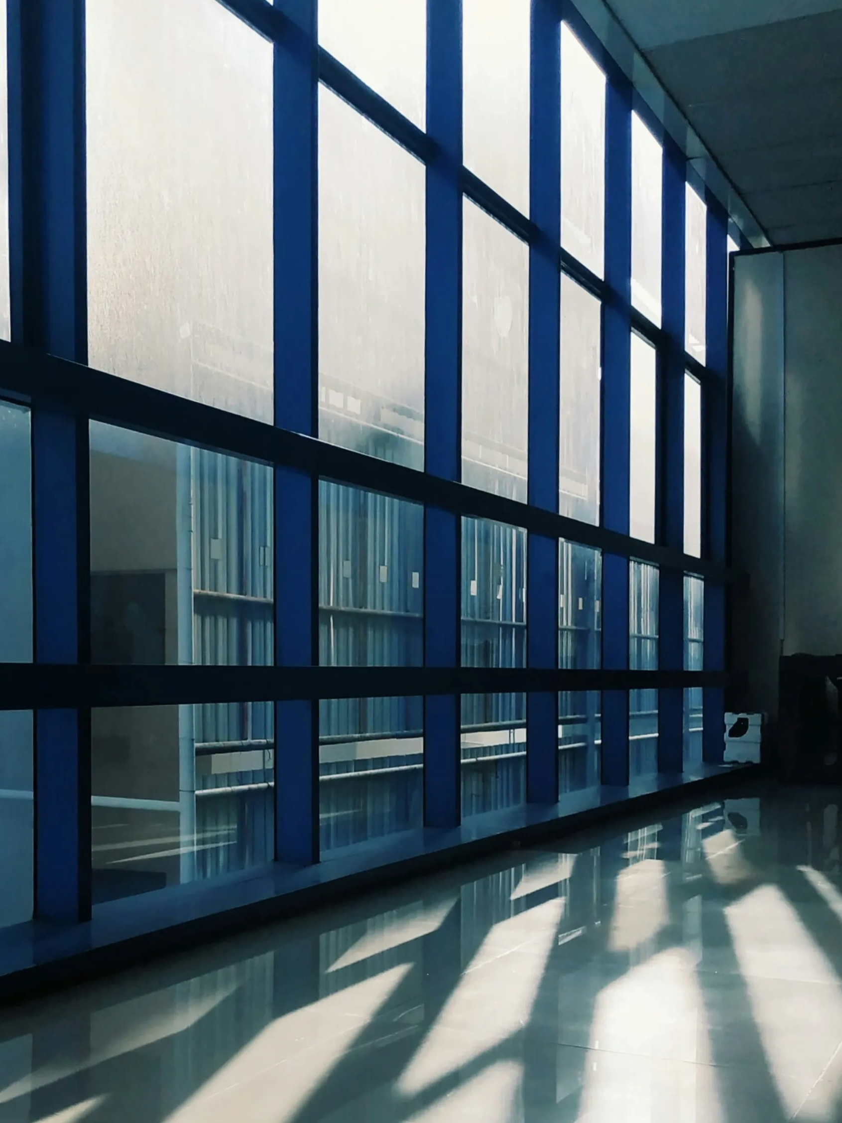 Blue-framed glass corridor with sunlight casting long shadows on the floor