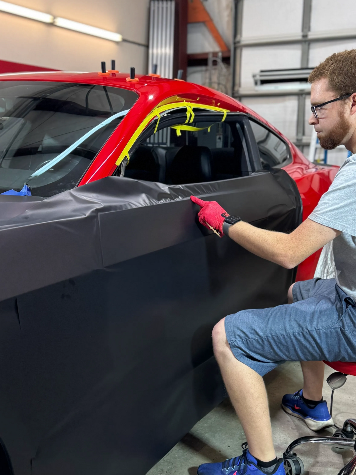 Man applying black vinyl wrap to a red car door in a garage