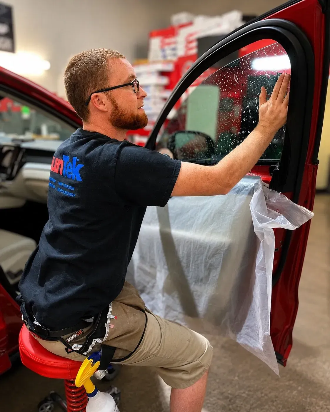 Man installing window tint on a red car door in a workshop