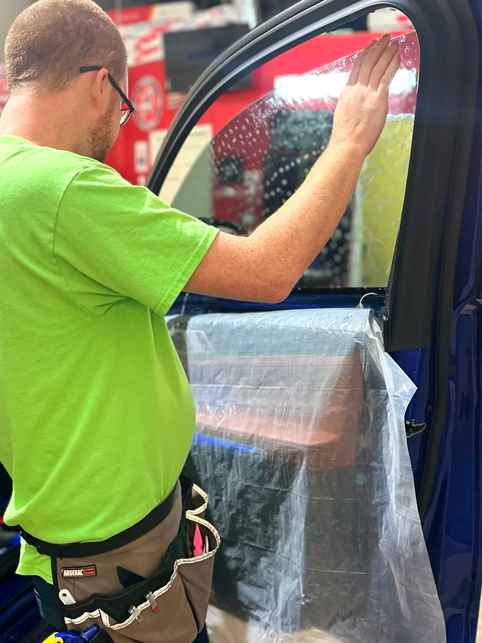Worker in a neon green shirt installing a window film on a vehicle door.