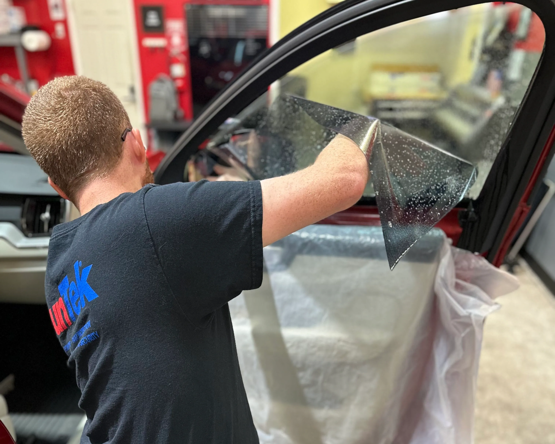 Person applying tint film to a car window with a squeegee inside a workshop.