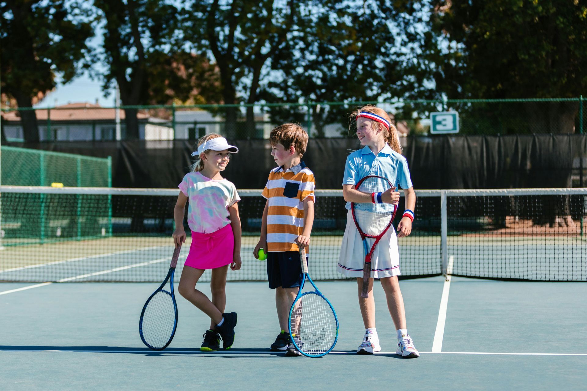 Three children on a tennis court, holding rackets and smiling. Blue court, green net, sunny day.