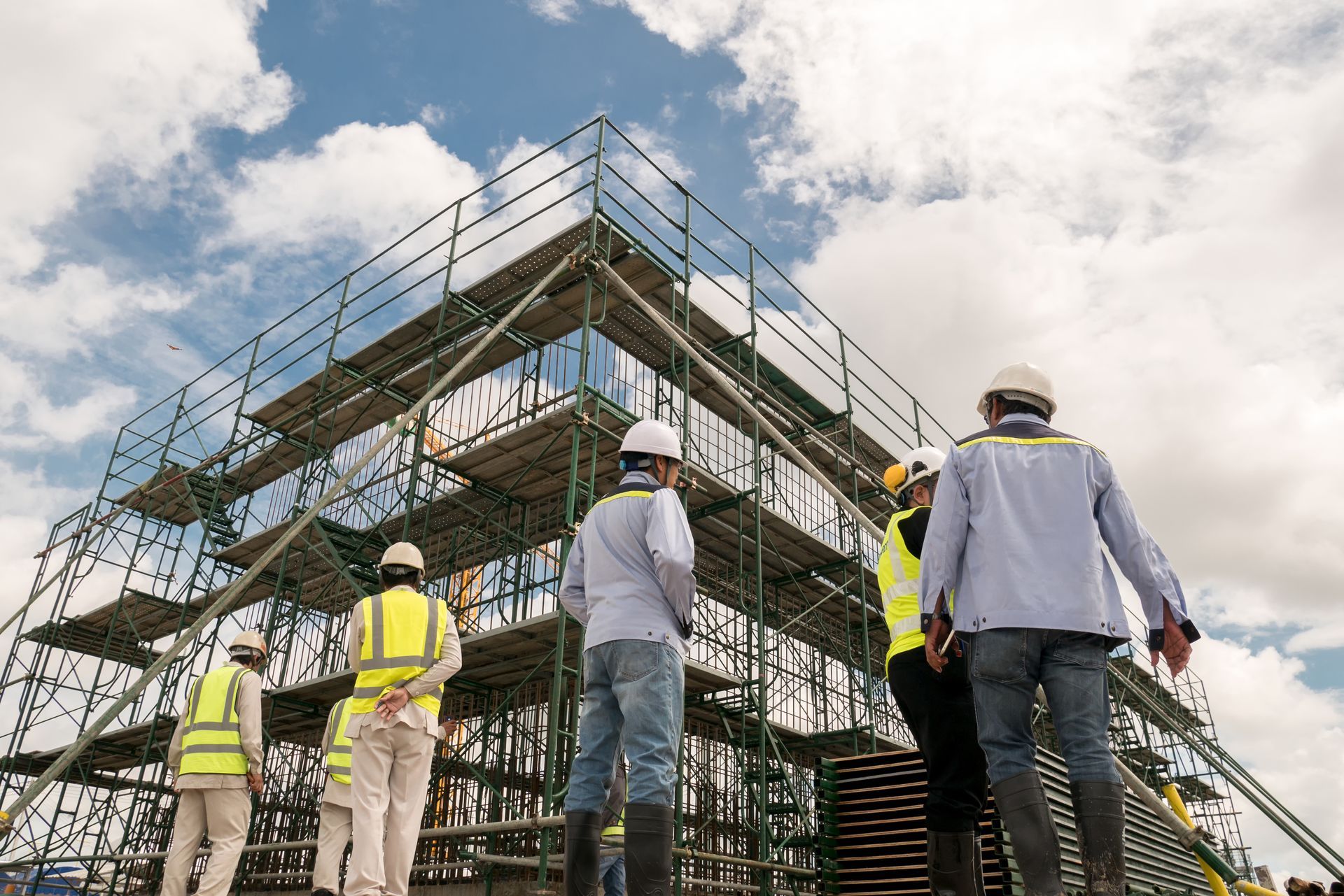 Construction workers in safety vests and hard hats surveying scaffolding under a cloudy sky.