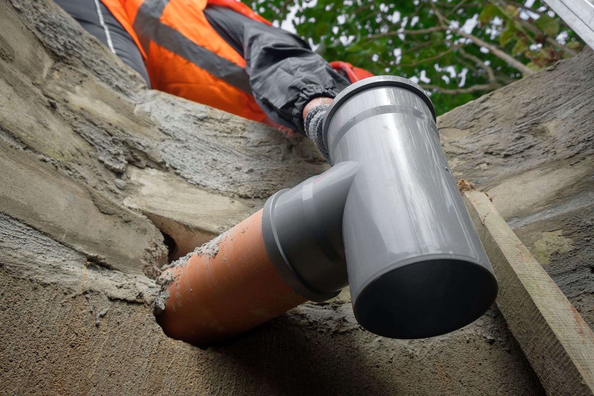 A person in an orange high-visibility jacket installs a grey plastic T-junction pipe into a hole in a concrete wall.