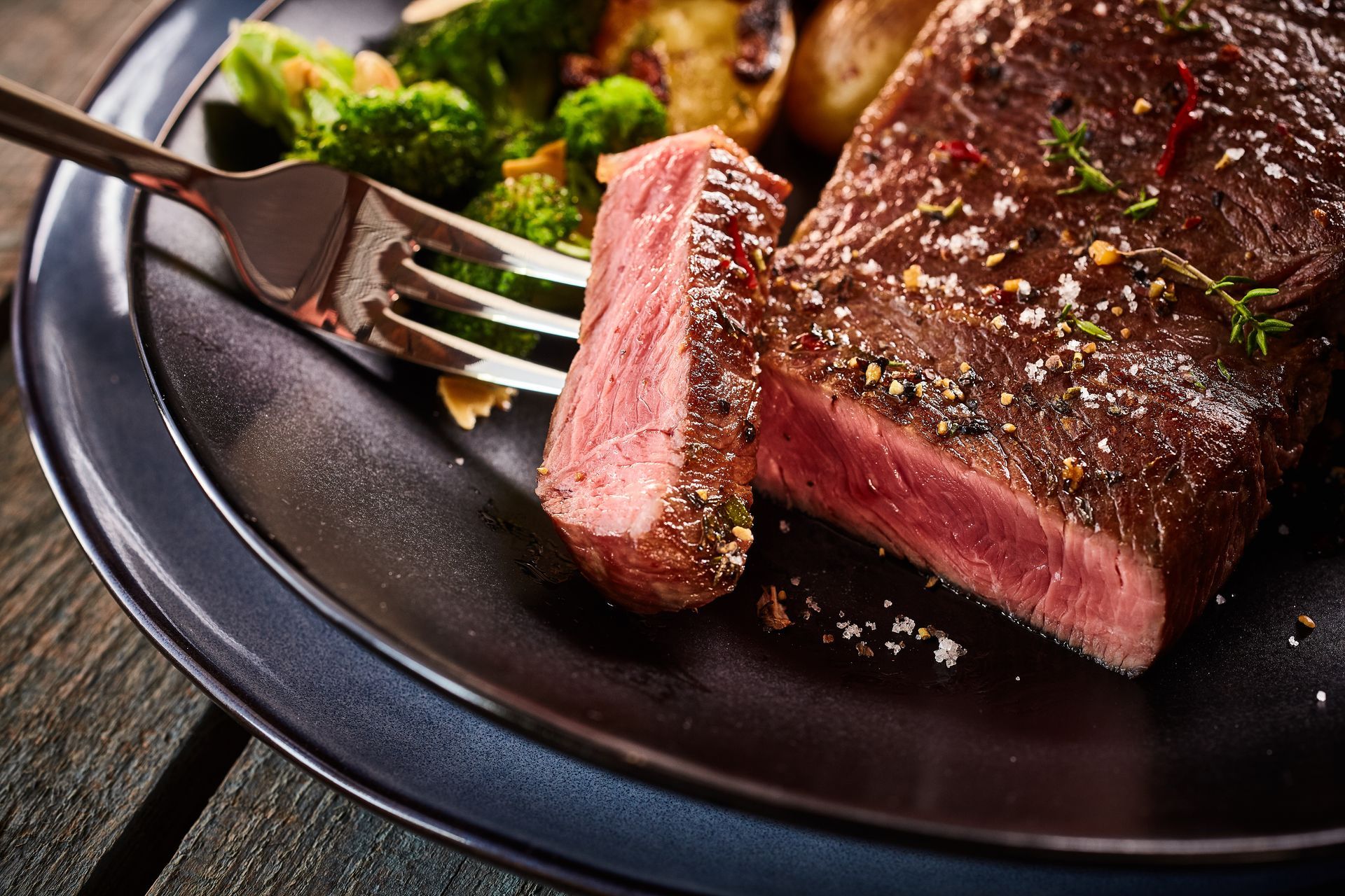 Rare steak on a black plate with a fork, garnished with greens and herbs