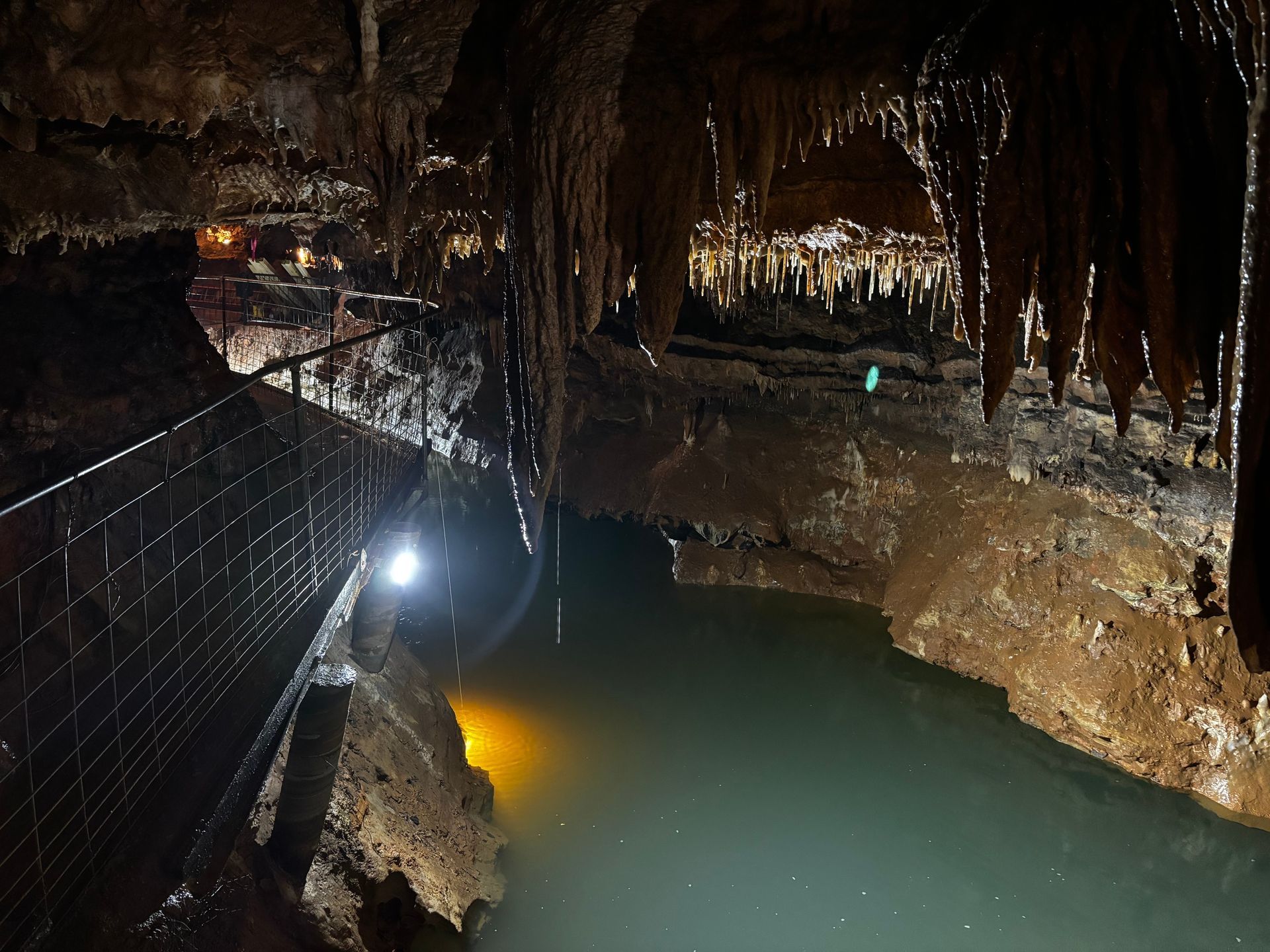 Dim cave pool with stalactites, a metal walkway, and a bright light reflecting on turquoise water.