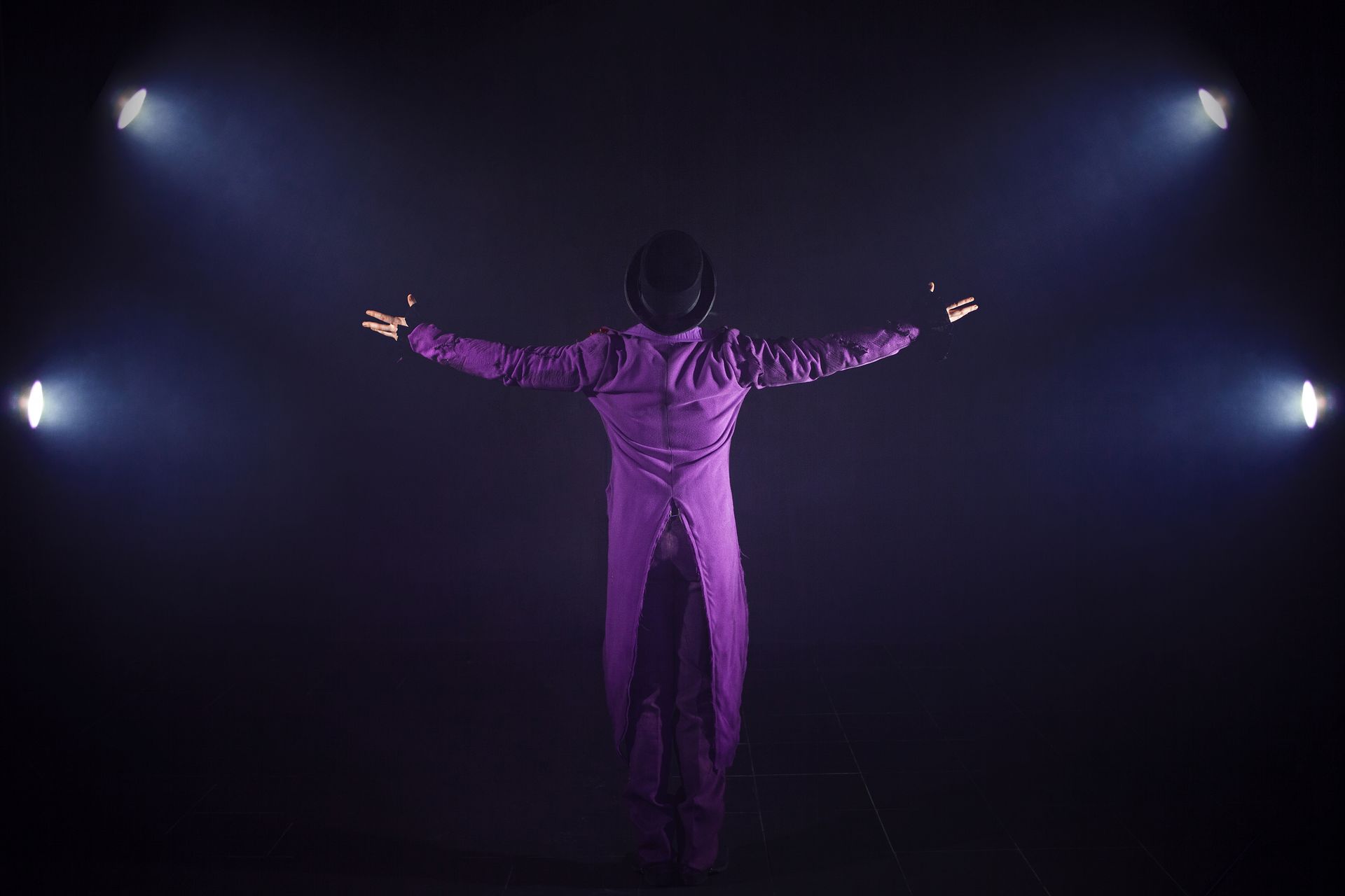 Person in purple suit stands center stage with arms outstretched under dramatic spotlights.