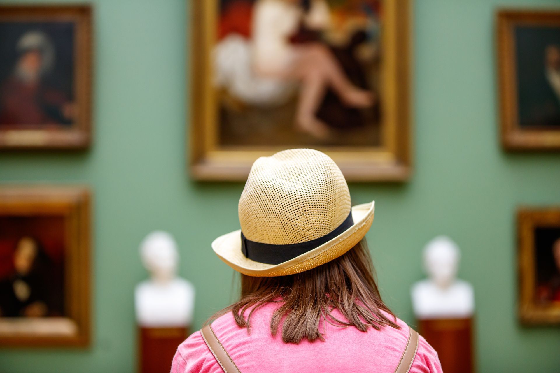 Visitor in straw hat and pink top viewing framed paintings in an art gallery