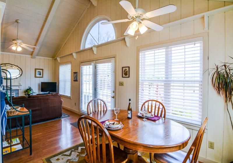 Bright dining area with round wooden table, high ceiling, ceiling fan, and large windows with blinds.