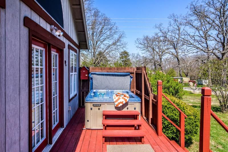Red wooden deck beside a house, with a hot tub and grassy yard under a bright sky.