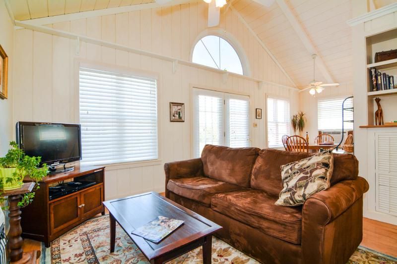 Bright living room with vaulted ceiling, brown sofa, TV, coffee table, and white shutters