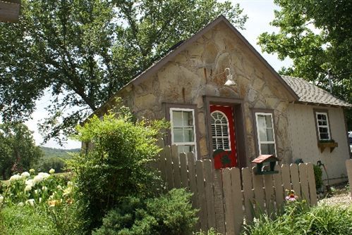 Small stone cottage with a red door, white windows, and a picket fence surrounded by greenery.