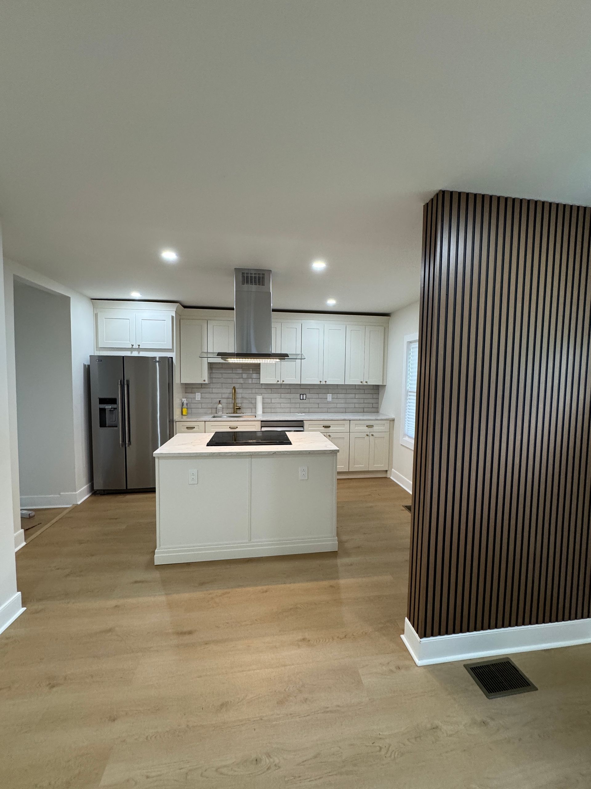 Modern open kitchen with white cabinets, island, stainless fridge, and wood floors, viewed from the living area