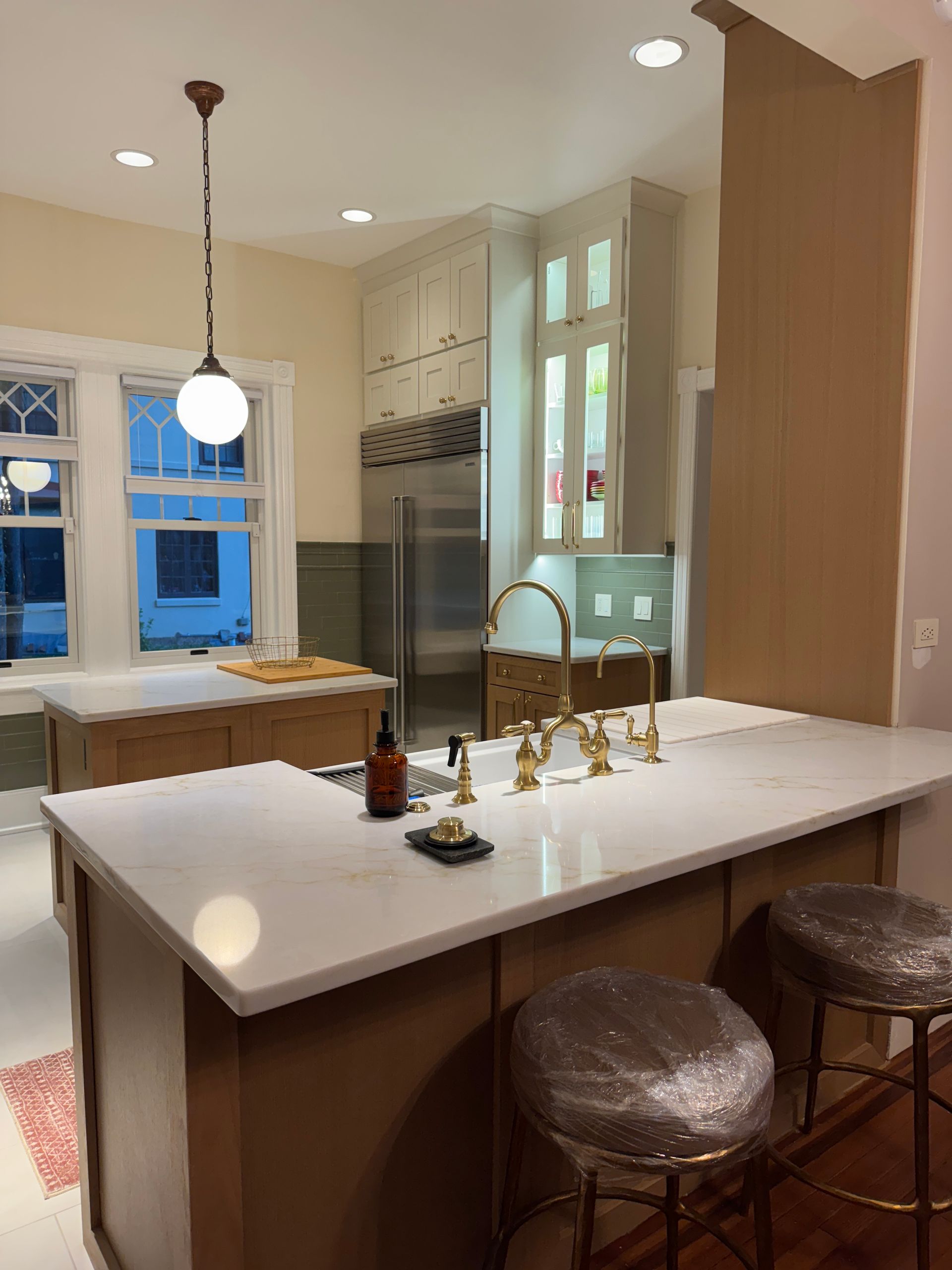 Bright kitchen with white island, pendant lights, wood cabinets, and two stools by a window.