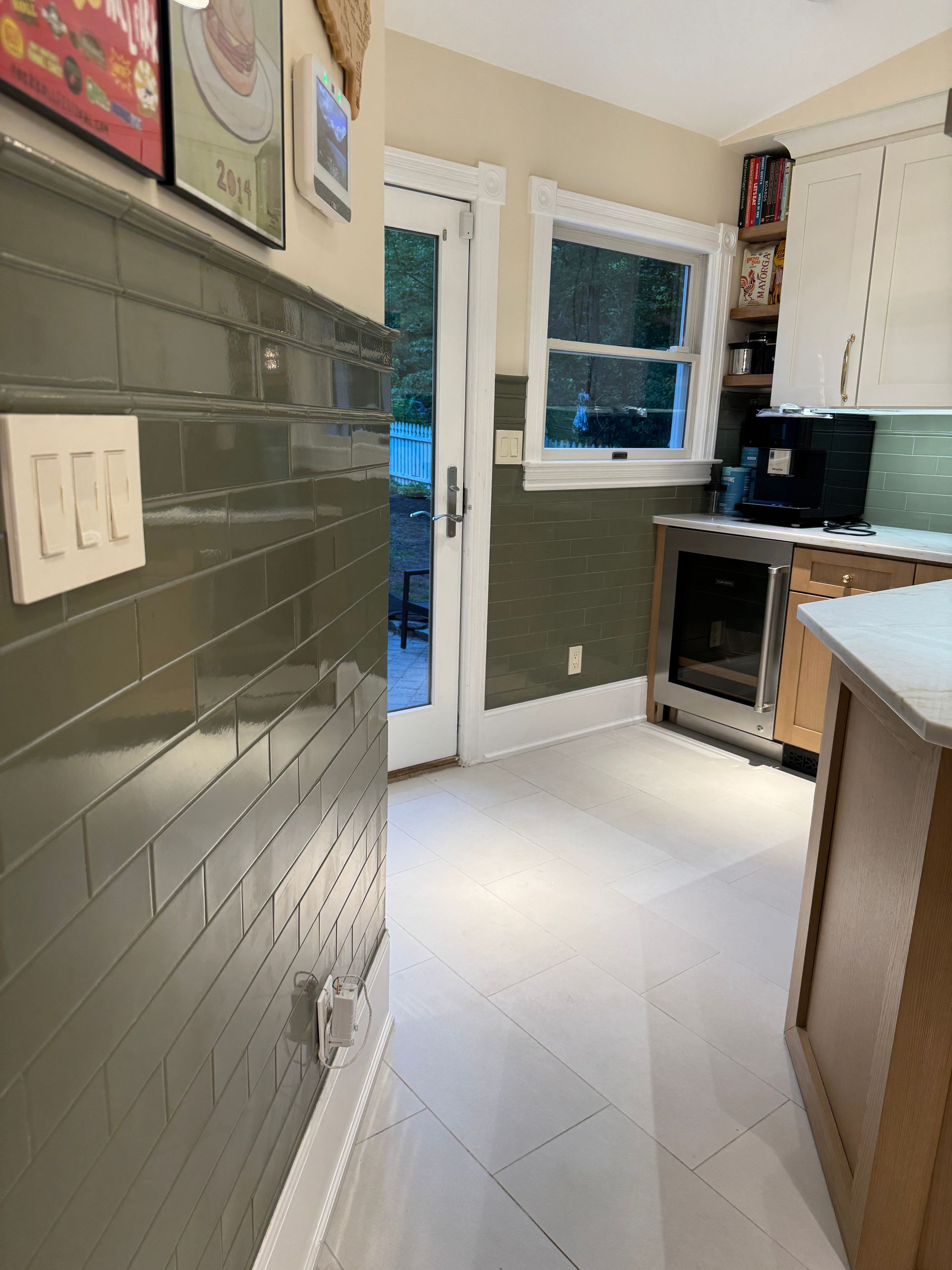 Narrow kitchen with gray tile walls, white cabinets, window, doorway, and countertop appliances.