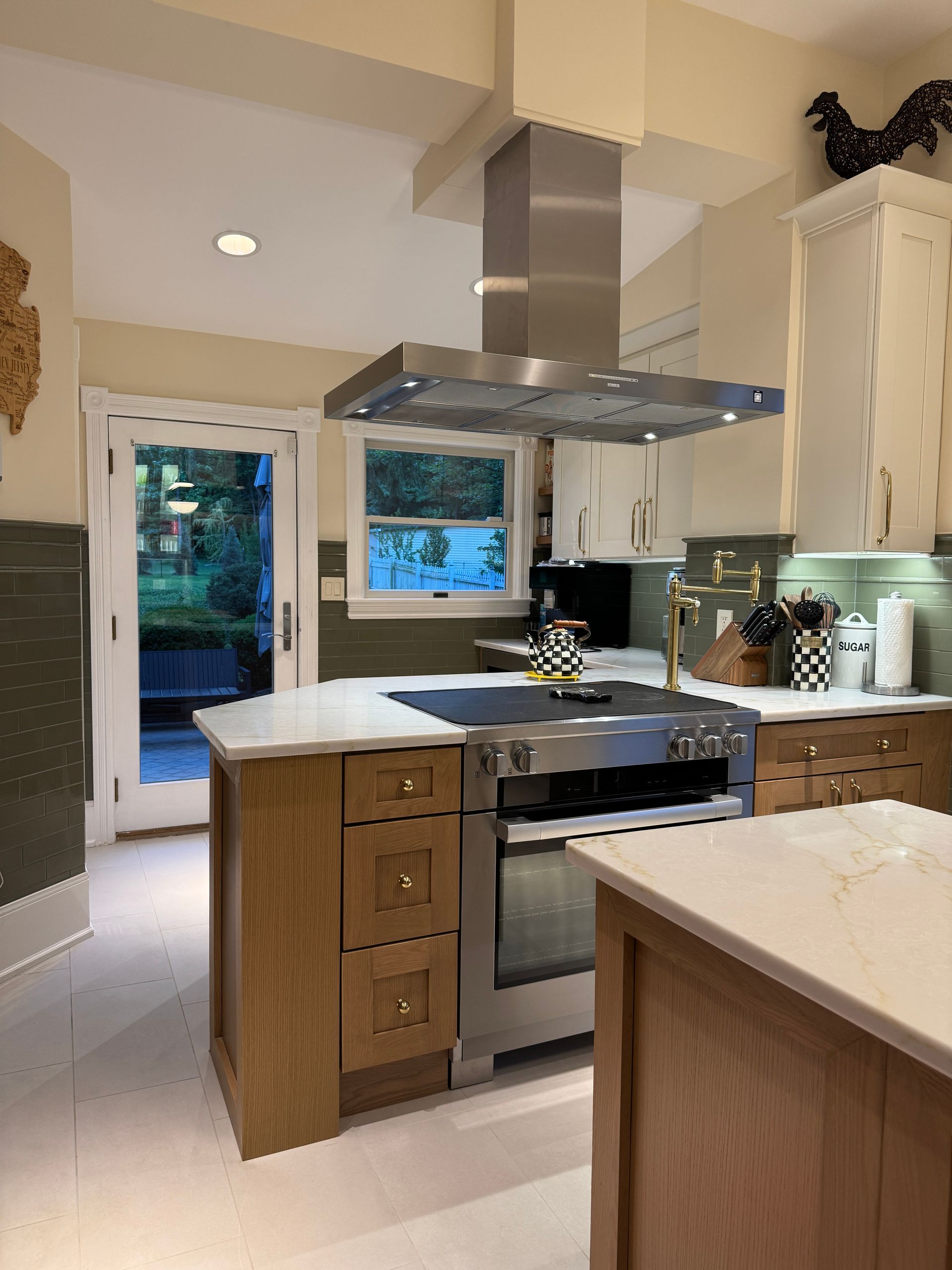 Modern kitchen with stainless steel range hood, island, wood cabinets, and a view of a backyard through an open door.