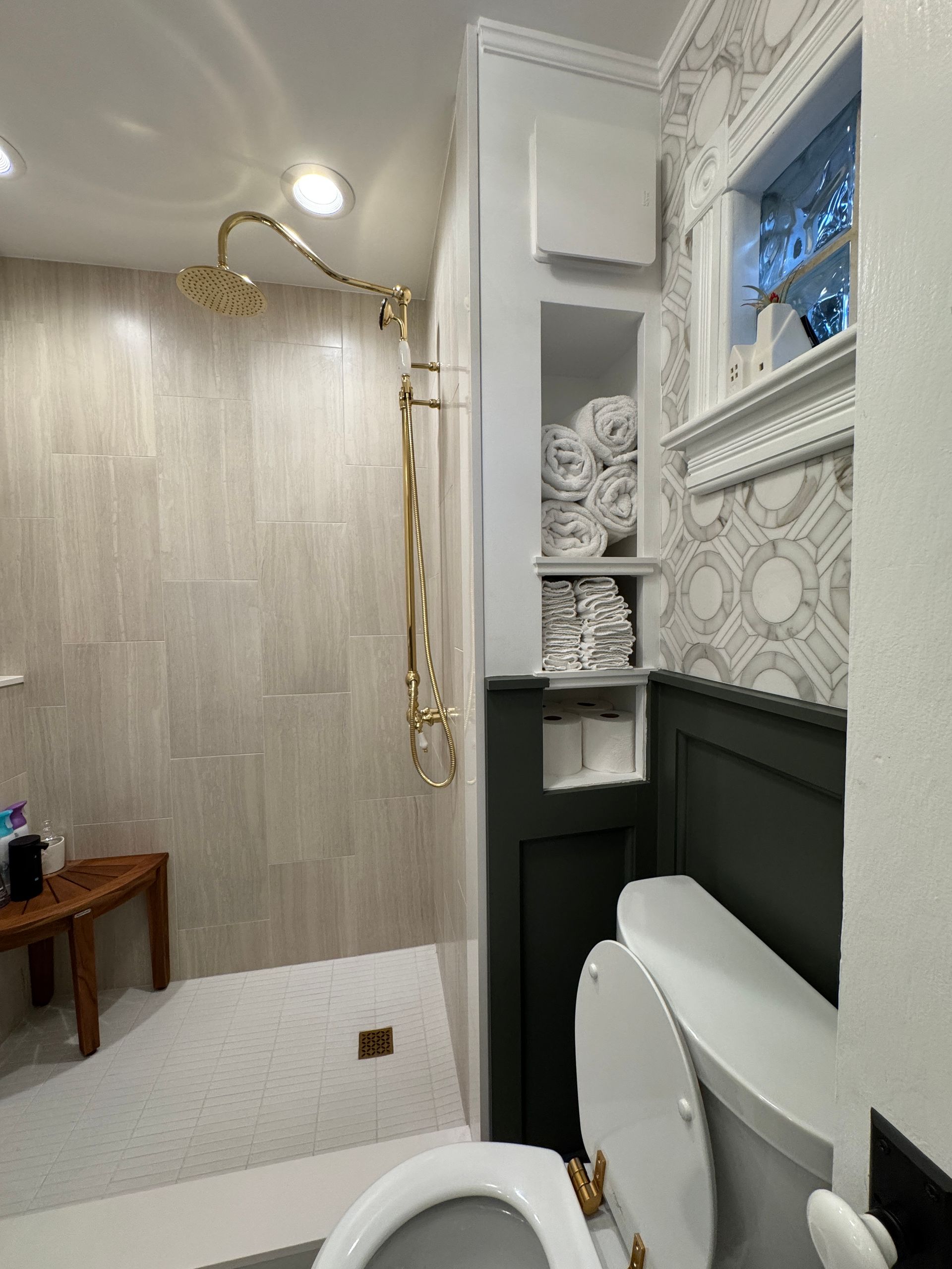 Bathroom with walk-in shower, white tile, gold shower fixture, wallpapered wall, and toilet in foreground