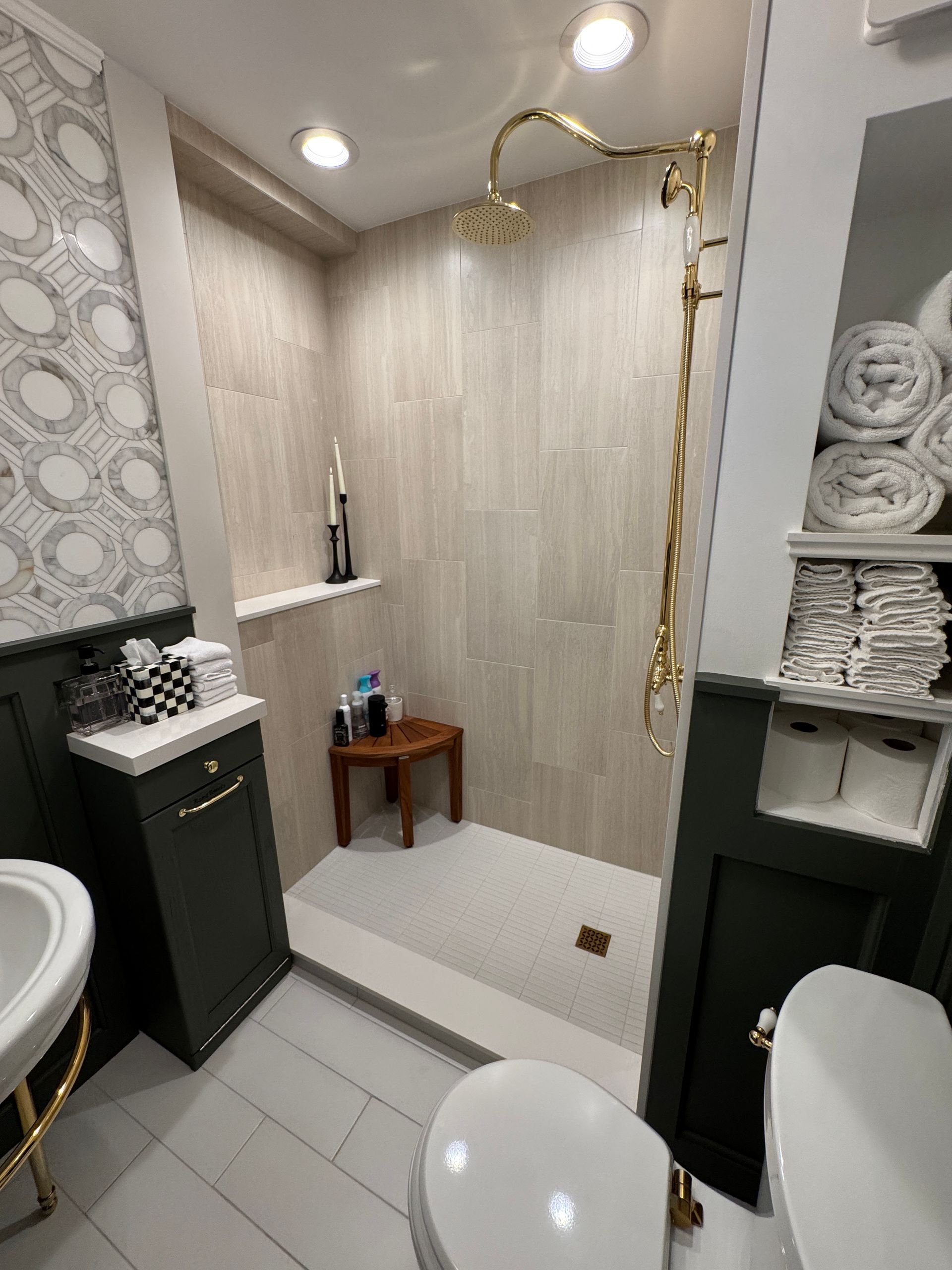 Small tiled bathroom with walk-in shower, gold fixtures, green cabinets, and stacked towels.