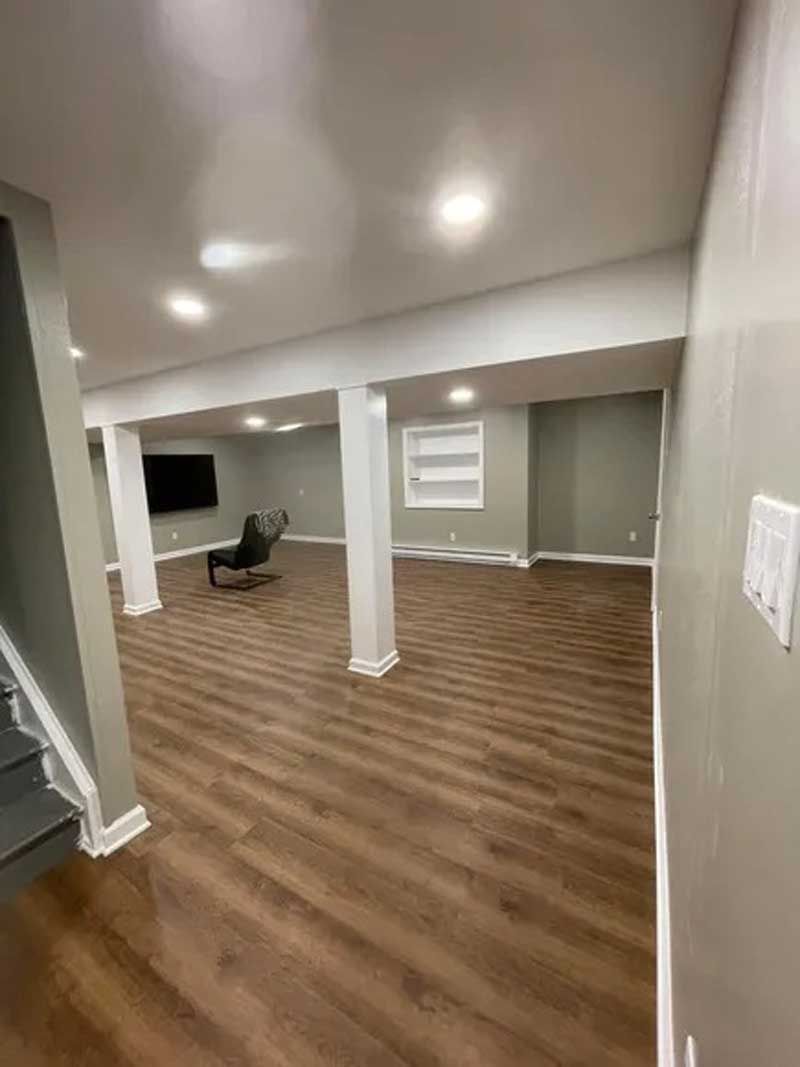 Empty finished basement with wood floors, gray walls, white columns, recessed lights, and a chair near the back wall.
