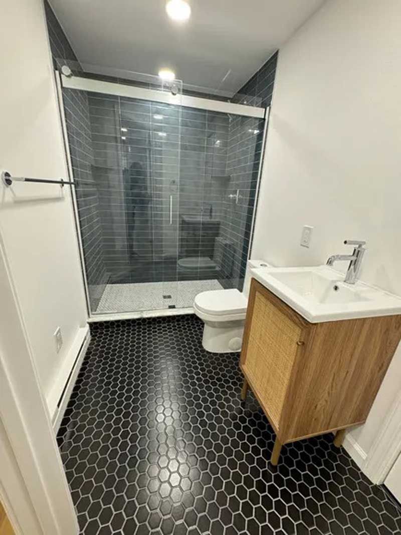 Bathroom with black hexagon tile floor, glass shower, light wood vanity, and white toilet.