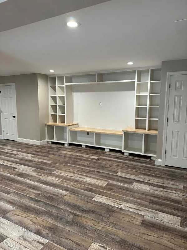 Built-in white shelves and cabinets on a wall, with a light wood bench. Dark wood-look floor.