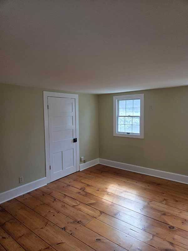 Empty room with wooden floor, white door and window, beige walls, and white trim.