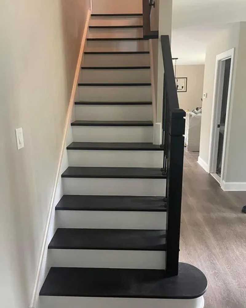 Staircase with black treads and white risers, black banister, beige walls, and wood-look flooring.