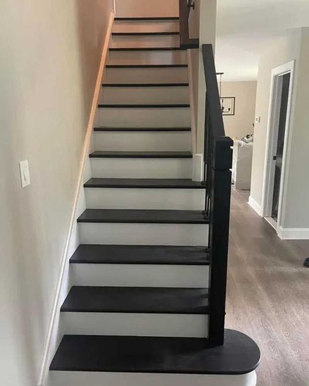 Staircase with black treads, white risers, and a black banister against a neutral-colored wall.