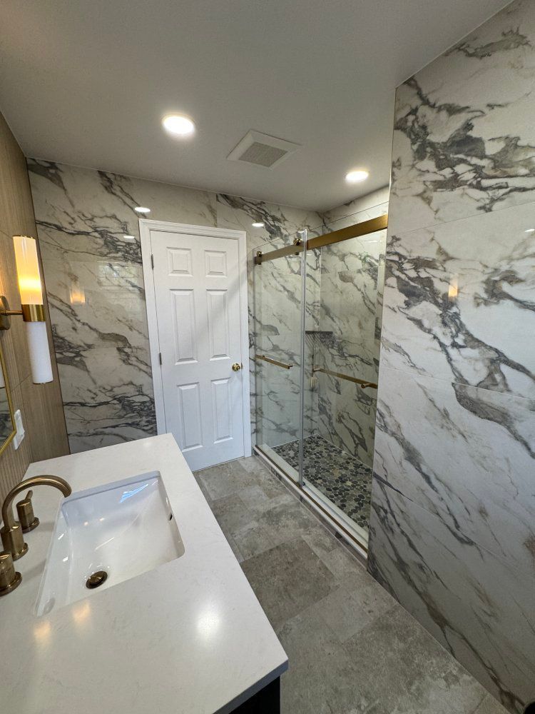 Modern bathroom with white and gray marble-look tile, glass shower, gold fixtures, white vanity, and gray floor tiles.