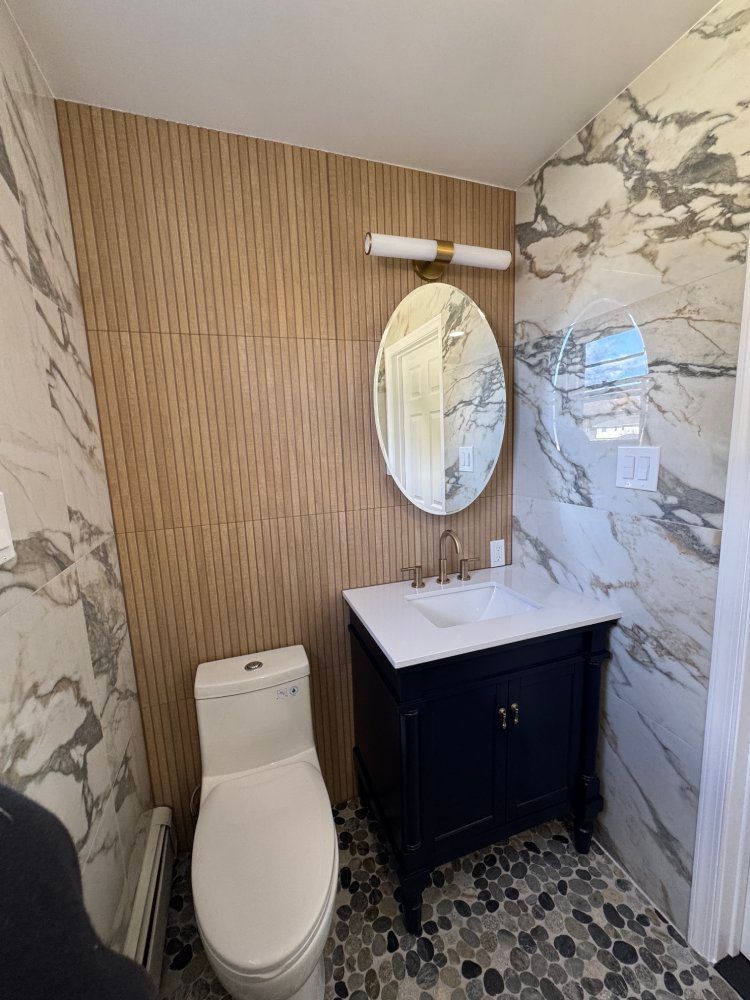 Small bathroom with wooden panel accent wall, marble tile wall, blue vanity, oval mirror, and pebble floor.
