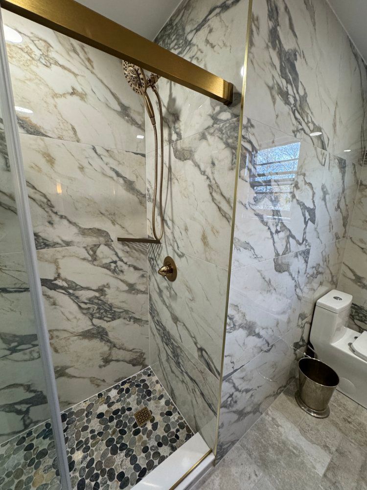 Marble-tiled shower with gold fixtures, glass door, and pebble floor. A toilet and trash can are also visible.