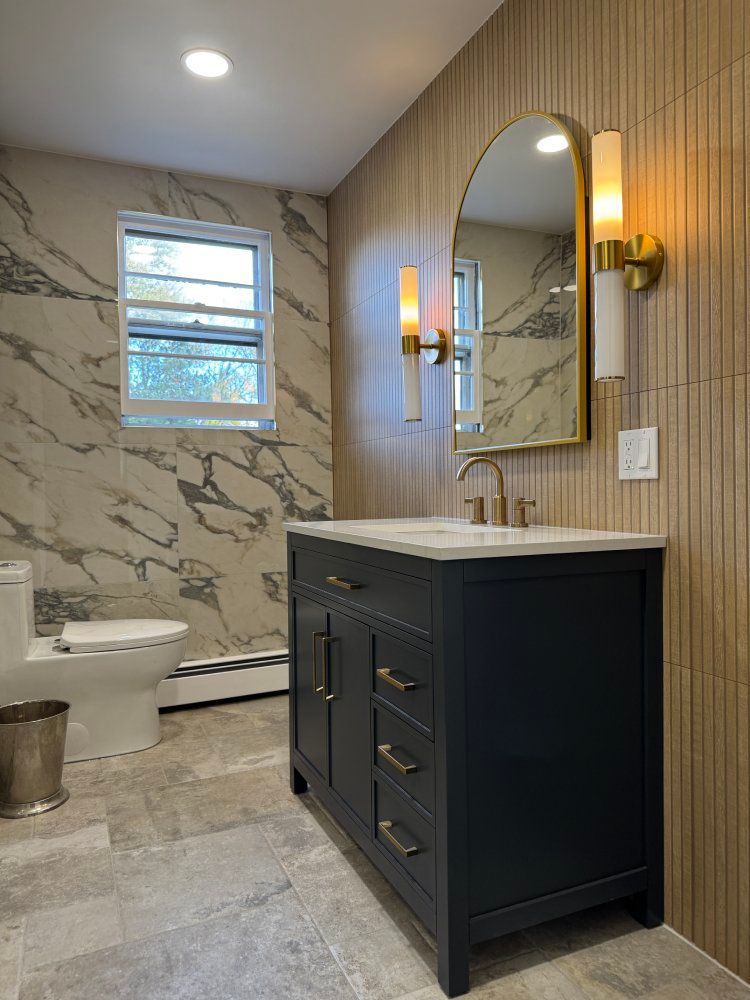 Bathroom with navy vanity, gold accents, marble and wood-look tile, and arched mirror.