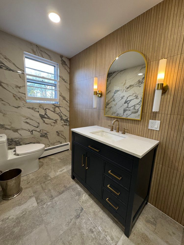 Modern bathroom with marble and wood-look walls, black vanity, gold accents, toilet, and window.