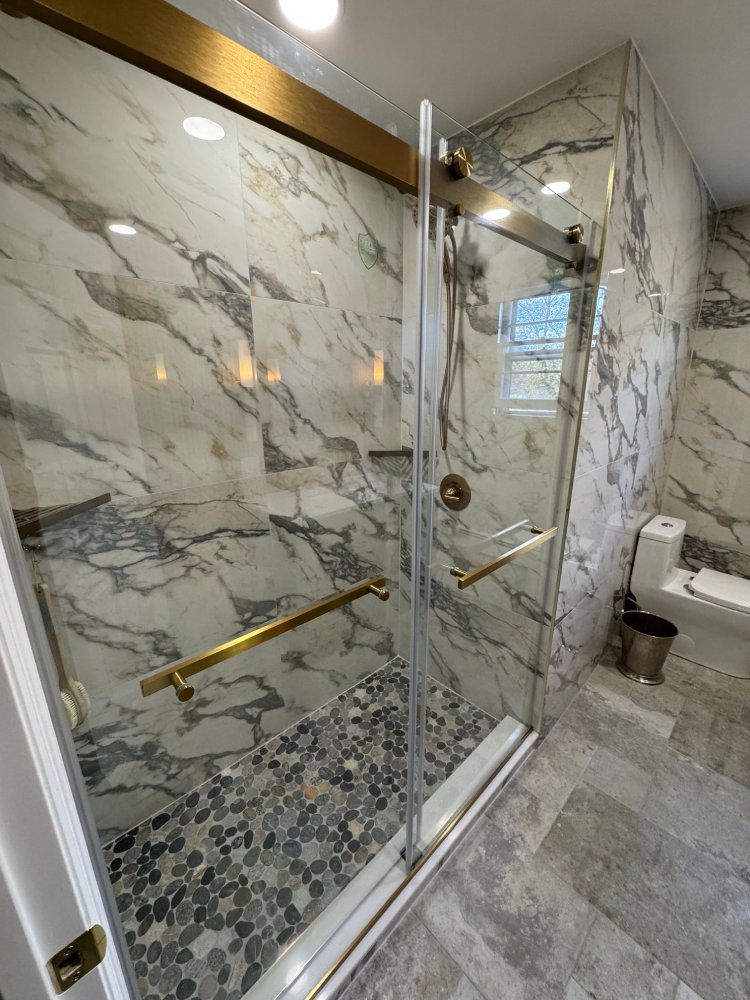 Marble-tiled shower with a clear sliding glass door, brass fixtures, and a pebble floor, in a modern bathroom.
