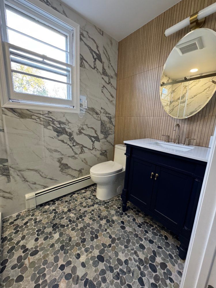 Bathroom with marble tile, blue vanity, pebble floor, and round mirror.
