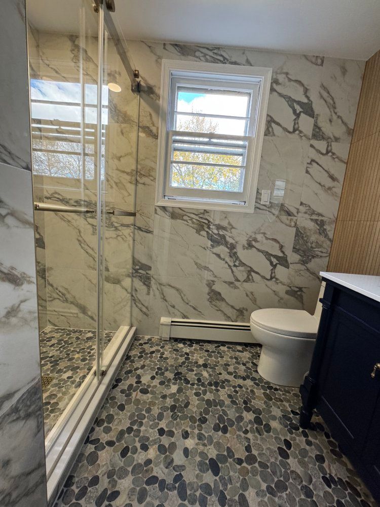 Bathroom with marble-patterned walls, pebble floor, glass shower, toilet, window, and navy vanity.