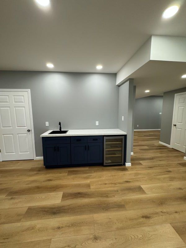 Blue bar cabinet with sink and wine cooler, white countertop, on wood floor, gray walls, recessed lighting.