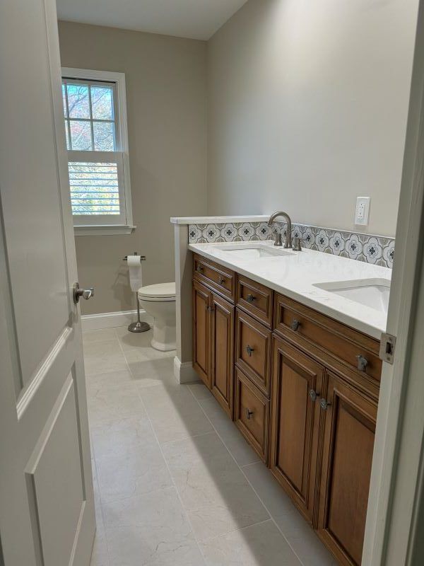 Bathroom with double sink vanity, toilet, and window. Brown cabinets, white countertops, beige walls, and light gray floor.