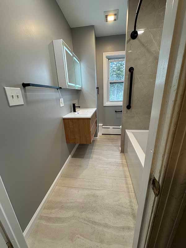 Narrow bathroom with light wood vanity, gray walls, tub, and tiled floors.