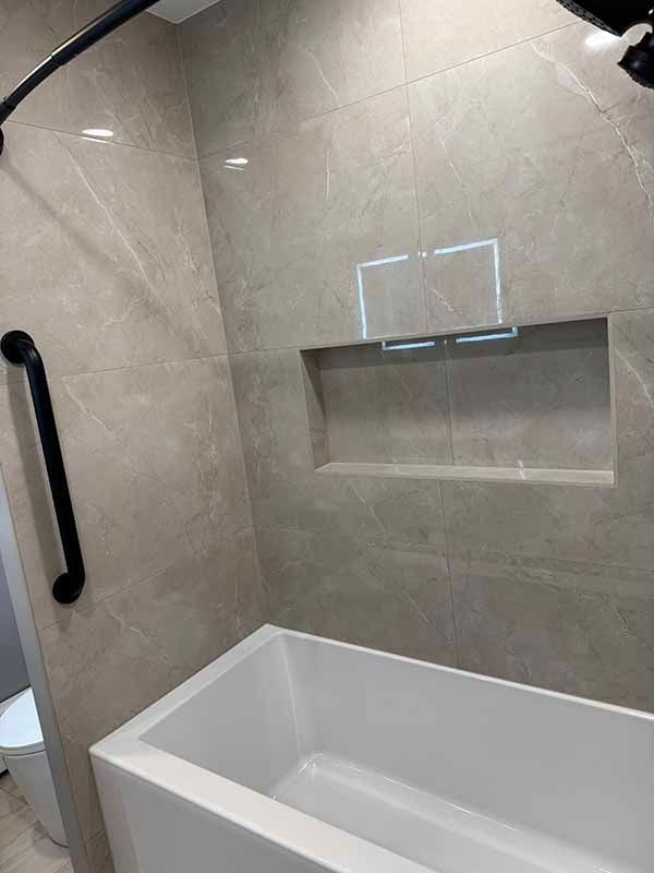 Bathroom with gray tiled walls, built-in shelf, white tub, black grab bar, and shower fixture.