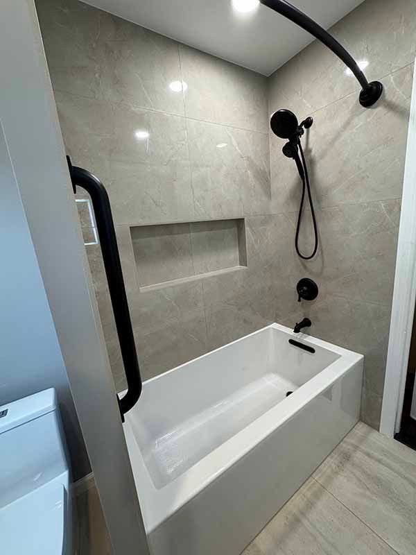 Modern bathroom with a white rectangular tub, gray tiled walls, and black shower fixtures.